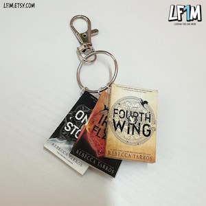 Może przedstawiać: Brelok z miniaturowymi zawieszkami w kształcie książek. Zawieszki zawierają tytuły "ON A WING", "IRON FLAME" i "FOURTH WING" autorstwa Rebeccy Yarros. Brelok ma srebrne zapięcie i kółko trzymające zawieszki.