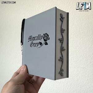 Könnte beinhalten: Ein graues Buch mit dem Schriftzug "Morally Grey" und einer schwarzen Rose. Das Buch hat einen schwarzen Riemen und ein dekoratives Rankenmuster auf dem Buchrücken. Das Buch wird von einer Hand gehalten.