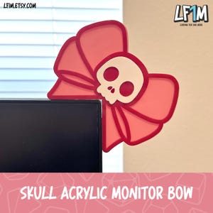 May include: A red acrylic monitor bow with a skull design. The bow is positioned on the corner of a black monitor. The skull is off-white with red eye sockets. The text "SKULL ACRYLIC MONITOR BOW" is at the bottom.