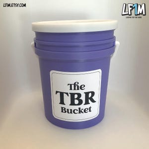 Könnte beinhalten: Ein lila Eimer mit weißem Deckel und Griff. Auf dem Eimer befindet sich ein weißes Etikett mit dem schwarzen Text "The TBR Bucket". Ideal zum Organisieren von Büchern, eine praktische Aufbewahrungslösung.