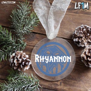 May include: A clear, round Christmas ornament with the name "Rhyannon" in white script. The ornament has a blue design and is hanging from a white ribbon. The background includes pine needles and pine cones.