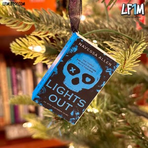 May include: A small book-shaped Christmas tree ornament with the title "LIGHTS OUT" and author "NAVESSA ALLEN". The cover displays a blue skull on a black background. The ornament is suspended from a black ribbon and hangs on a Christmas tree.