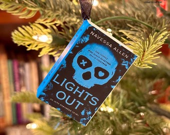 May include: A small book-shaped Christmas tree ornament with the title "LIGHTS OUT" and author "NAVESSA ALLEN". The cover displays a blue skull on a black background. The ornament is suspended from a black ribbon and hangs on a Christmas tree.