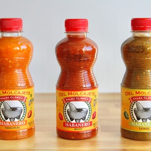 May include: Three bottles of Del Molcajete habanero salsa in original, rojo, and verde flavors. Each bottle has a red cap and a label with a mortar and pestle graphic. The original is orange, rojo is red, and verde is green.