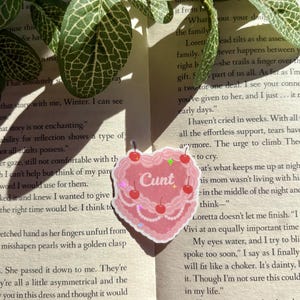 May include: A pink heart-shaped sticker with the word "Cunt" in the centre, decorated with cherries and glitter. The sticker is placed on a book page with green leaves in the background.