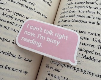 Può includere: Un segnalibro rosa a forma di fumetto con il testo "I can't talk right now. I'm busy reading." è posto su un libro aperto. Le pagine del libro sono ingiallite e il testo è visibile.