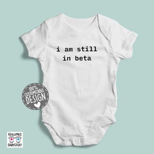 May include: White baby onesie with black text that reads "i am still in beta".