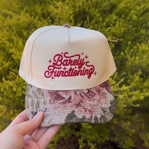 May include: A cream-colored baseball cap featuring the embroidered text "Barely Functioning" in a dark pink cursive font with small star accents. The brim of the cap is covered in a woven fabric with a detailed floral pattern in shades of pink, green, and tan.