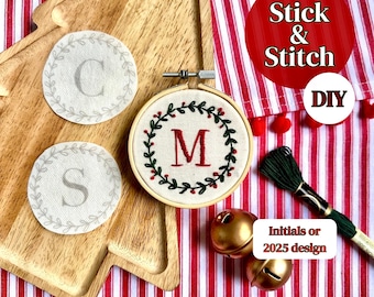 May include: A wooden tray holds three embroidered items: two round patches with letters "C" and "S", and a circular embroidery hoop with the letter "M". The hoop is decorated with a green wreath and red berries. The text "Stick & Stitch DIY" is visible.