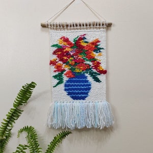 Puede incluir: Un tapiz de pared tejido con un vibrante arreglo floral en un jarr&oacute;n azul. Las flores son rojas, naranjas y amarillas, sobre un fondo blanco. El jarr&oacute;n tiene un dise&ntilde;o a rayas. La parte inferior tiene flecos y cuelga de una varilla de madera.