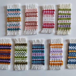 May include: Ten small woven wall hangings with fringe. The hangings are in various colors and patterns, including red, white, blue, green, purple, and orange.