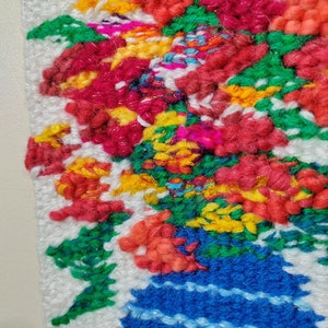 Puede incluir: Primer plano de una colorida y texturizada obra de arte textil. La obra representa un vibrante ramo de flores en un jarr&oacute;n azul. Las flores son una mezcla de rojo, amarillo, naranja y verde. El jarr&oacute;n tiene rayas azules horizontales. El fondo es blanco.