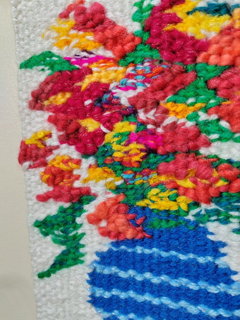 Puede incluir: Primer plano de una colorida y texturizada obra de arte textil. La obra representa un vibrante ramo de flores en un jarr&oacute;n azul. Las flores son una mezcla de rojo, amarillo, naranja y verde. El jarr&oacute;n tiene rayas azules horizontales. El fondo es blanco.