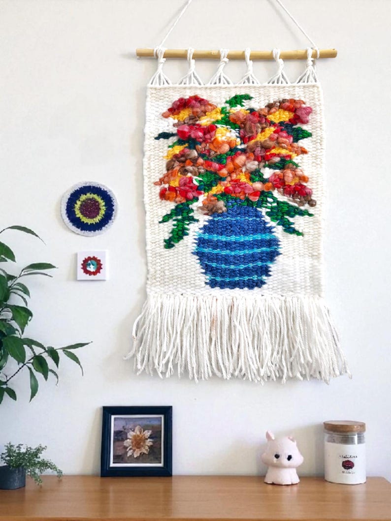 Puede incluir: Un tapiz de pared tejido que presenta un jarr&oacute;n azul lleno de flores coloridas en tonos rojos, naranjas y amarillos. La obra de arte est&aacute; hecha de hilo blanco con una franja en la parte inferior. Tambi&eacute;n se ve una peque&ntilde;a imagen enmarcada y otros objetos decorativos.