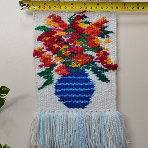 Puede incluir: Tapiz de pared tejido a mano con un dise&ntilde;o floral. Muestra un jarr&oacute;n azul lleno de flores rojas, naranjas y amarillas. El tapiz mide aproximadamente 15 cm de ancho y tiene una franja en la parte inferior.