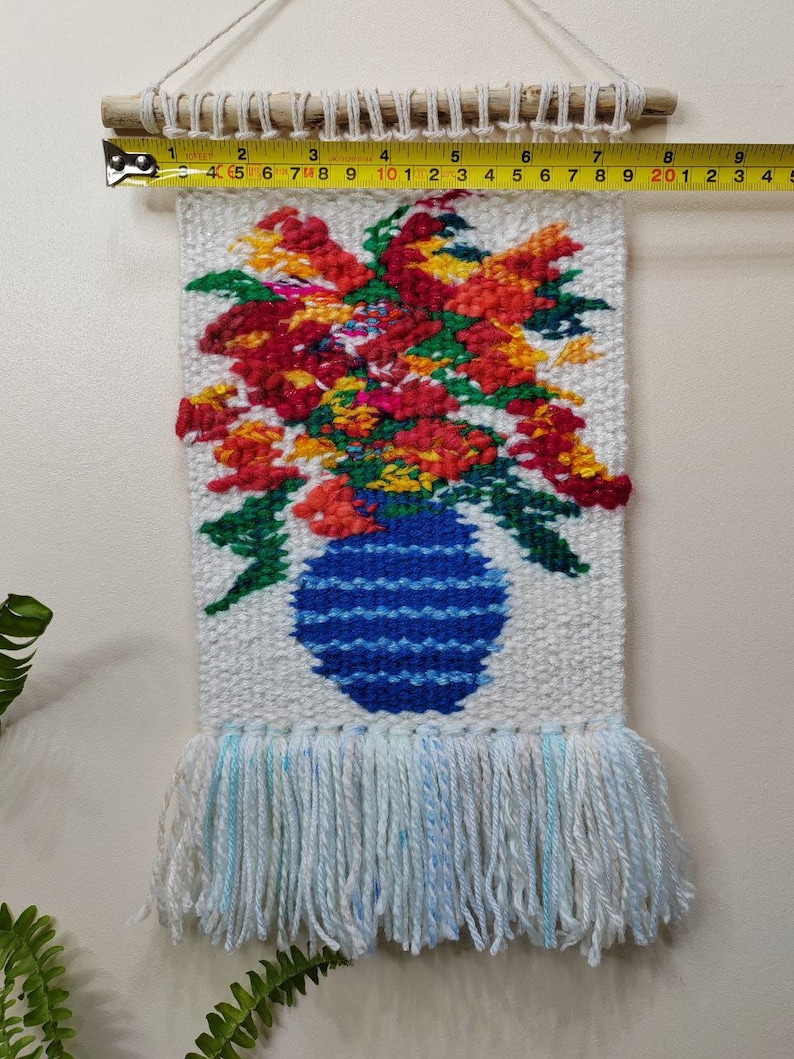 Puede incluir: Tapiz de pared tejido a mano con un dise&ntilde;o floral. Muestra un jarr&oacute;n azul lleno de flores rojas, naranjas y amarillas. El tapiz mide aproximadamente 15 cm de ancho y tiene una franja en la parte inferior.