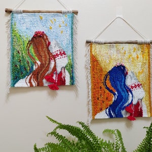 May include: Two woven wall hangings with a woman in a white dress and red sash. The woman on the left has brown hair and the woman on the right has blue hair. Both hangings have a white fringe.