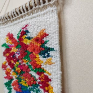 Puede incluir: Un tapiz de pared tejido con un vibrante arreglo floral en un jarr&oacute;n azul. Las flores son una mezcla de rojo, naranja, amarillo y rosa, sobre un fondo blanco. La pieza est&aacute; montada en una varilla de madera con un cord&oacute;n de fibra natural para colgar.