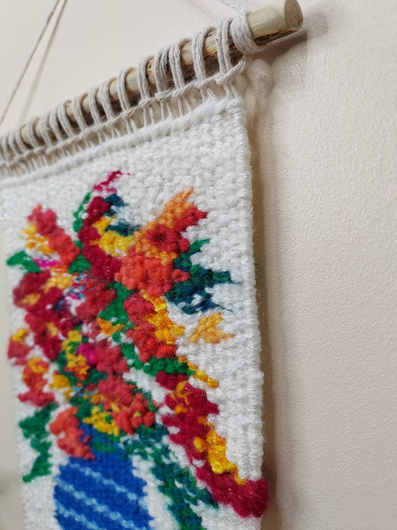 Puede incluir: Un tapiz de pared tejido con un vibrante arreglo floral en un jarr&oacute;n azul. Las flores son una mezcla de rojo, naranja, amarillo y rosa, sobre un fondo blanco. La pieza est&aacute; montada en una varilla de madera con un cord&oacute;n de fibra natural para colgar.