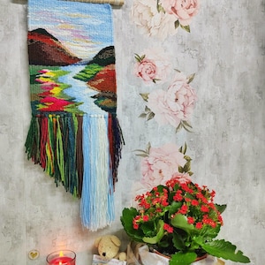 May include: A woven wall hanging with a landscape scene featuring a river, mountains, and a blue sky. The hanging has colorful fringe at the bottom. A small teddy bear, a potted plant with red flowers, and a candle are in the foreground.