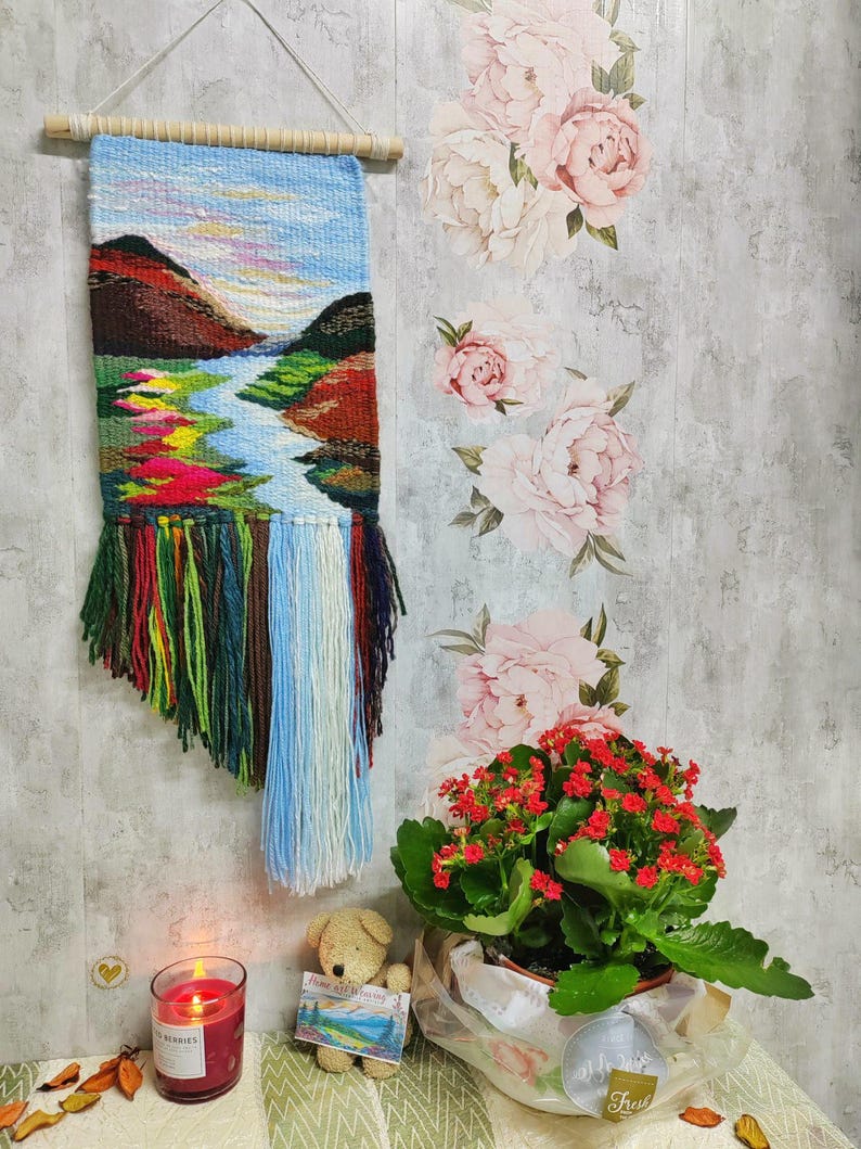 May include: A woven wall hanging with a landscape scene featuring a river, mountains, and a blue sky. The hanging has colorful fringe at the bottom. A small teddy bear, a potted plant with red flowers, and a candle are in the foreground.