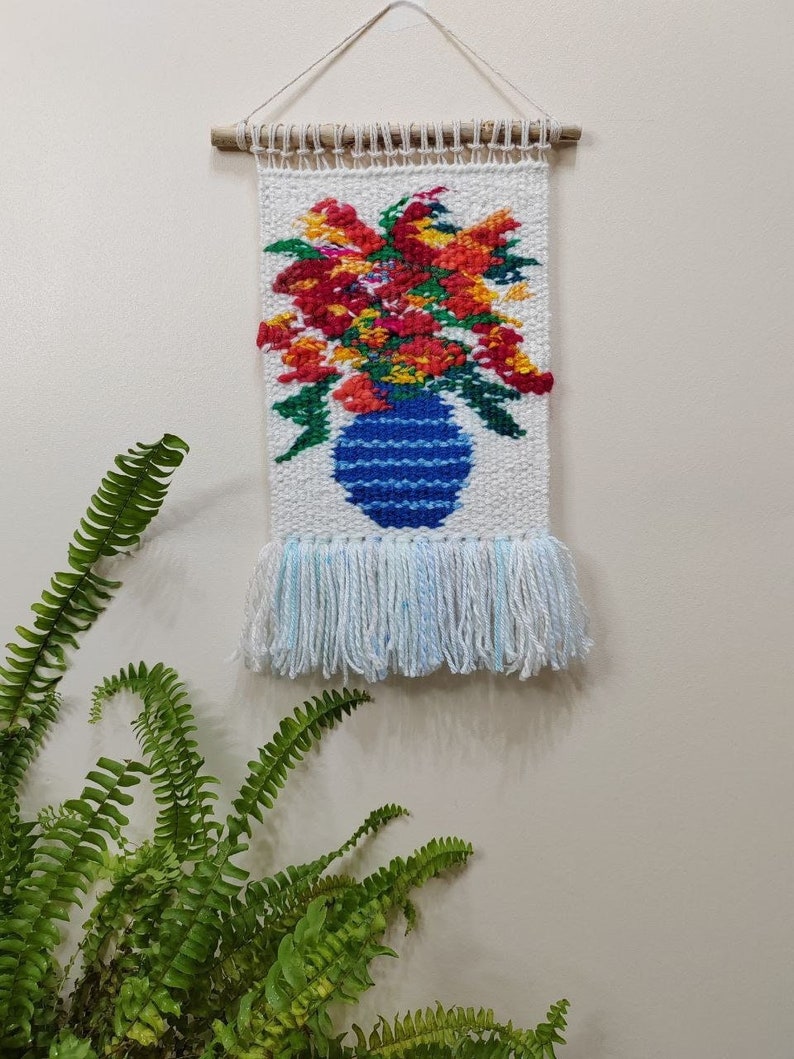 Puede incluir: Un tapiz de pared tejido con un vibrante arreglo floral en un jarr&oacute;n azul. Las flores son rojas, naranjas y amarillas, sobre un fondo blanco. El jarr&oacute;n tiene un dise&ntilde;o a rayas. La parte inferior tiene flecos y cuelga de una varilla de madera.