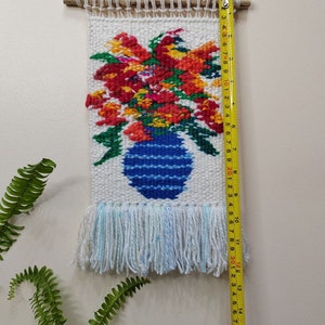 Puede incluir: Tapiz de pared tejido con un dise&ntilde;o floral en un jarr&oacute;n azul. El tapiz, de unos 40 cm de largo, muestra flores rojas, naranjas y amarillas. Tiene una franja en la parte inferior y est&aacute; colgado de una varilla de madera.