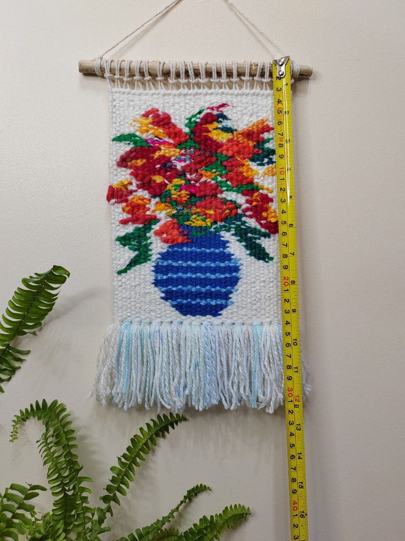 Puede incluir: Tapiz de pared tejido con un dise&ntilde;o floral en un jarr&oacute;n azul. El tapiz, de unos 40 cm de largo, muestra flores rojas, naranjas y amarillas. Tiene una franja en la parte inferior y est&aacute; colgado de una varilla de madera.