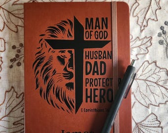Può includere: Un diario marrone con le parole "MAN OF GOD, HUSBAND, DAD, PROTECTOR, HERO" e un disegno di testa di leone. Il nome "James" è in basso. Una penna nera è appoggiata sul diario. È presente anche il testo "1 Corinzi 16".