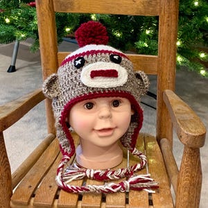 May include: A brown and white crocheted monkey hat with a red pom-pom on top. The hat has a white stripe and a red nose. The hat is on a wooden rocking chair.