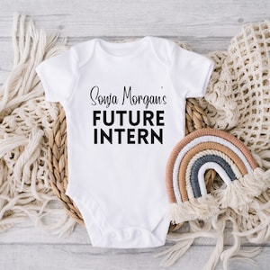 May include: White baby onesie with black text that reads "Sonja Morgan's Future Intern". The onesie is lying on a woven basket with a rainbow-colored macrame hanging next to it.