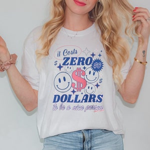 Peut inclure: T-shirt blanc avec un motif coloré indiquant "It Costs Zero Dollars to be a nice person". Le design comprend des smileys, des symboles de dollars et des étoiles roses, bleues et blanches. Un vêtement décontracté pour tous les jours.