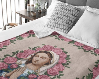 May include: A beige blanket with a religious image encircled by pink roses. The image shows a woman in a blue robe and white veil with hands clasped in prayer. The blanket is on a bed with white pillows.