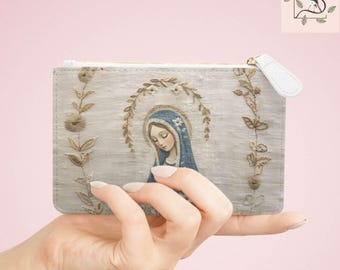 May include: A rectangular clutch with a zipper closure, featuring an illustration of a woman with a blue veil and halo. The clutch is light gray with gold floral embroidery and a white zipper pull. The image is held by a hand.