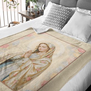May include: A beige throw blanket featuring a religious image of a woman with a halo, wearing a light blue and floral patterned shawl. The blanket has a brown border and is displayed on a bed with white bedding.