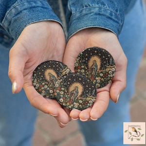 Puede incluir: Tres imanes decorativos redondos y negros con una imagen religiosa de una mujer con halo, rodeada de flores y estrellas. Los imanes se sostienen en manos abiertas sobre un fondo de chaqueta vaquera.