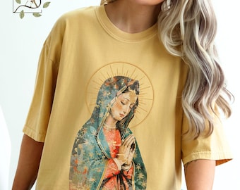 以下が含まれることがあります： マスタードイエローのTシャツ。ヴィンテージスタイルのグラフィックで、青と緑のベールをかぶり、祈りの手を合わせ、赤い衣服を着た宗教的な人物が描かれています。画像には光輪があります。Tシャツは半袖で、クルーネックです。