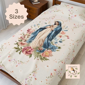 May include: A cream-colored bed blanket featuring a central illustration of the Virgin Mary wearing a blue mantle, surrounded by a floral arrangement of pink, blue, and white flowers. The blanket includes a small logo with a bird and the text GoodVibes2You.