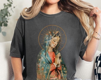 以下が含まれることがあります： 濃いグレーのTシャツに、青い金色の後光の中にいる聖母マリアのヴィンテージ風の画像がプリントされています。画像の下には「Behold Your Mother」という文字がプリントされています。
