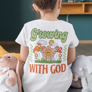 Op de afbeelding: Wit T-shirt met een groene "Growing with God" afbeelding. De afbeelding toont een glimlachende bloem met twee armen omhoog, omringd door paddenstoelen, wolken en een zon.