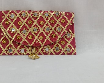 Puede incluir: Un bolso de mano rojo con solapa, adornado con patrones geométricos dorados y pequeños acentos de espejo reflectantes. El bolso de mano tiene una borla dorada y probablemente está hecho de tela.