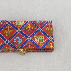 Può includere: Una scatola rettangolare con una vivace copertura in tessuto fantasia. Il design presenta colori rosso, blu e oro con motivi ripetuti di elefanti, paisley e forme geometriche. Una chiusura color oro assicura la scatola.