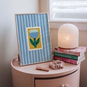 Puede incluir: Una impresi&oacute;n enmarcada con un dise&ntilde;o floral azul y amarillo, colocada sobre una mesita de noche rosa. Una l&aacute;mpara blanca, libros, una scrunchie y un l&aacute;piz labial tambi&eacute;n est&aacute;n en la mesita. La impresi&oacute;n tiene un fondo a rayas azules.