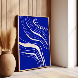May include: A framed abstract art print with a deep blue backdrop and cream-coloured, flowing lines. The artwork is presented in a light wood frame, leaning against a wooden wall. A decorative vase with dried plants is in the foreground.