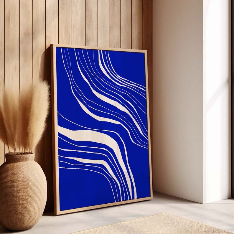 May include: A framed abstract art print with a deep blue backdrop and cream-coloured, flowing lines. The artwork is presented in a light wood frame, leaning against a wooden wall. A decorative vase with dried plants is in the foreground.