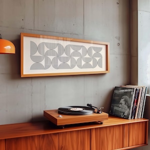 May include: A framed artwork with a geometric black and white pattern hangs above a wooden record player and a stack of vinyl records. The record player is on a wooden cabinet. An orange lamp hangs from the ceiling.
