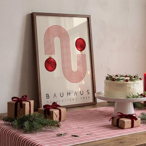 May include: A framed poster featuring red concentric lines forming an S-shape with two red ornaments, labeled BAUHAUS Ausstellung 1919. The print is displayed on a table with a striped runner, a frosted cake, and small gift boxes tied with red velvet ribbons.