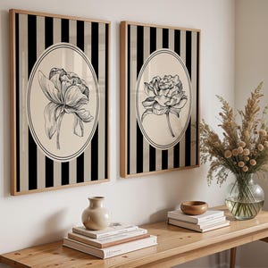 May include: Two framed wall art pieces. Each features a black and beige striped background with a beige oval containing a black and white floral illustration. The frames are light wood. The art is displayed on a white wall.