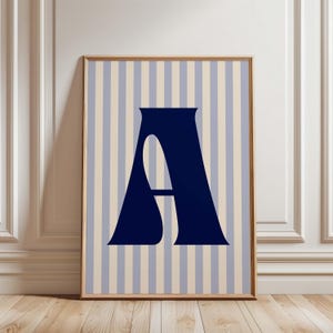 May include: Framed art print featuring a navy blue letter "A" against a striped background of light blue and beige vertical lines. The artwork is displayed in a wooden frame, leaning against a white wall and wooden floor.