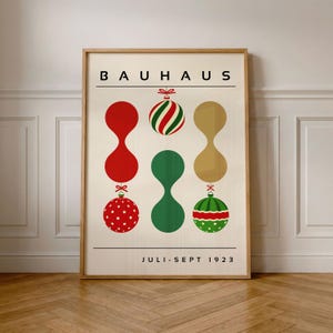 May include: A rectangular poster in a light wood frame featuring the text BAUHAUS at the top and JULI-SEPT 1923 at the bottom. The design displays six abstract shapes resembling Christmas ornaments in red, green, and gold, arranged in three vertical columns against a cream background.